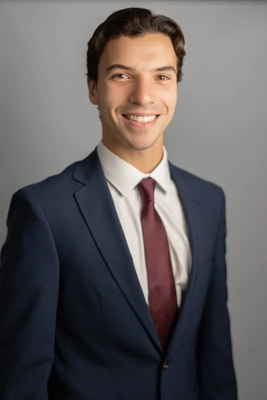 Marco smiles directly at the camera in a navy blazer, white dress shirt, and burgundy tie against a neutral gray background.