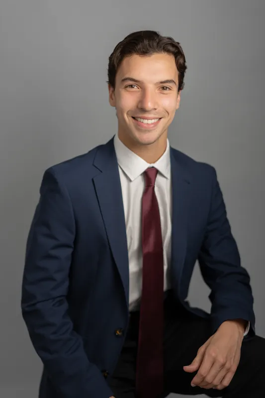 Marco stands with a relaxed smile, hand near his chest, wearing navy blazer and burgundy tie with soft gray backdrop.