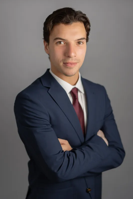 Marco faces the camera with arms crossed, wearing navy blazer and burgundy tie in a studio portrait setting.