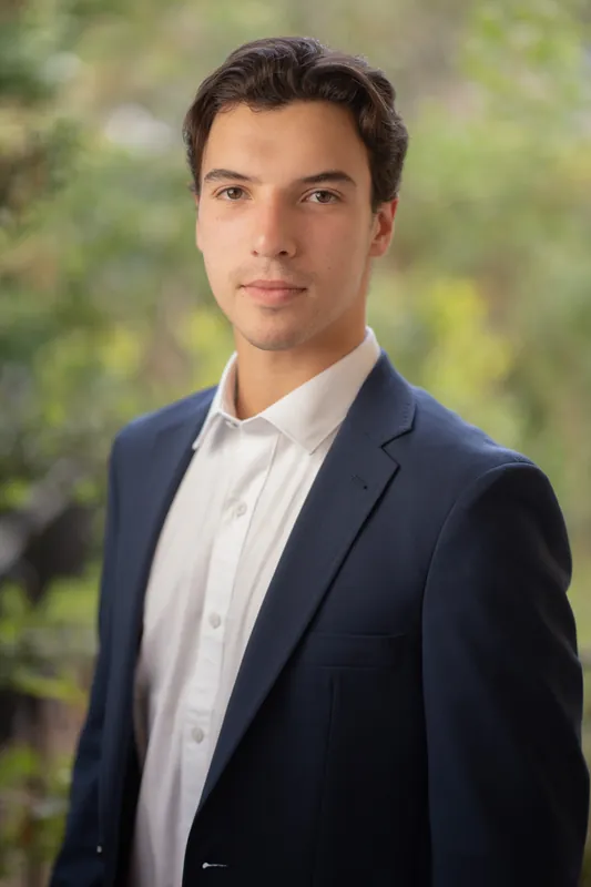 Marco faces the camera in a navy blazer and white shirt, with blurred foliage behind him.