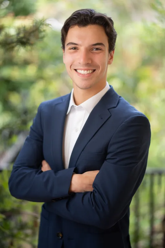 Marco smiles with arms crossed in navy blazer and white shirt, green garden setting visible in the background.