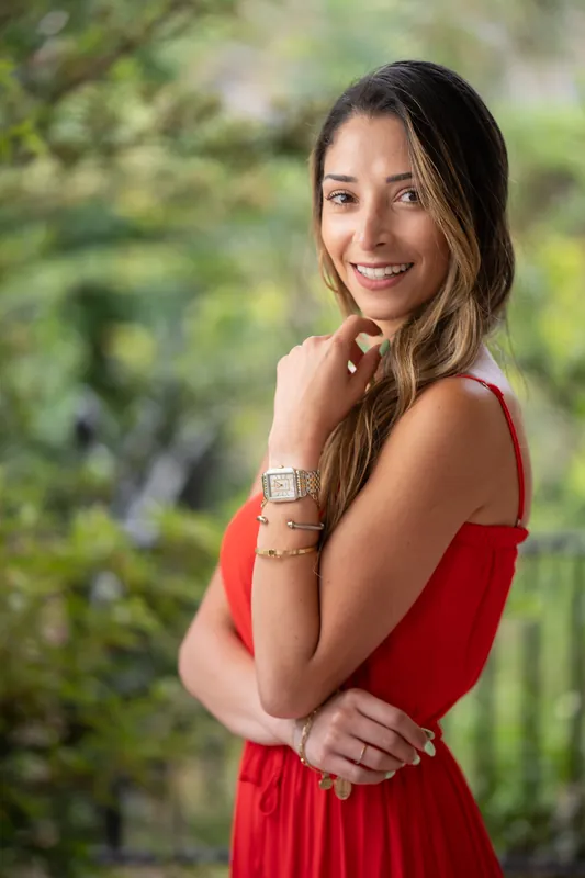 Natasha tilts her head and grins while touching her necklace, wearing a red dress in a garden setting.