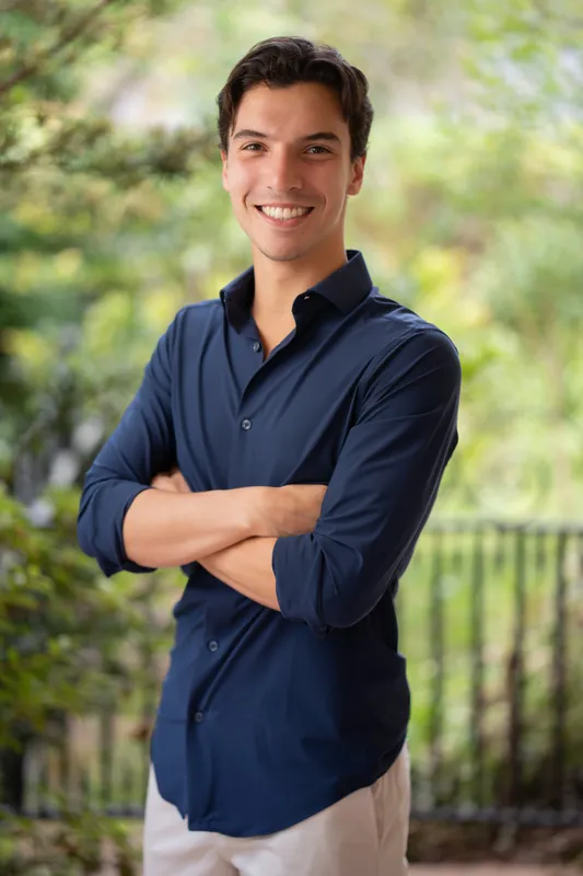 Marco smiles with arms crossed, wearing a navy button-up shirt and light pants against green landscaping.