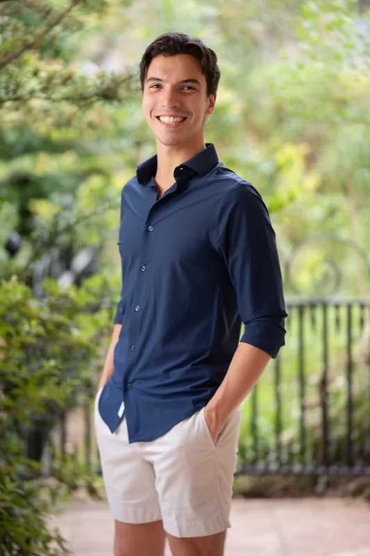 Marco stands casually in a navy polo and white shorts with hands in pockets, grinning outdoors.