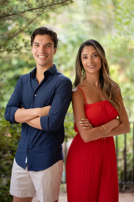 Natasha in red and Marco in navy pose together side-by-side, both smiling at Casa Futrell's leafy grounds.
