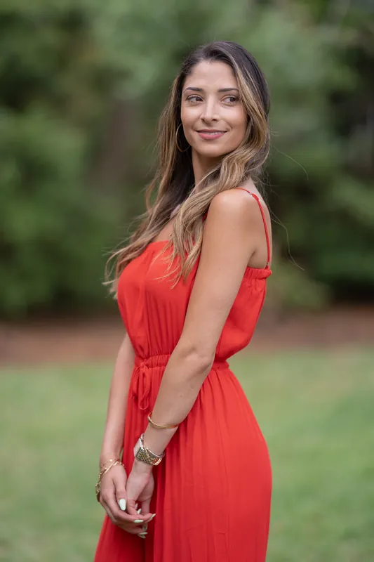 Natasha faces the camera over her shoulder in a red dress, with soft green foliage blurred in the background.