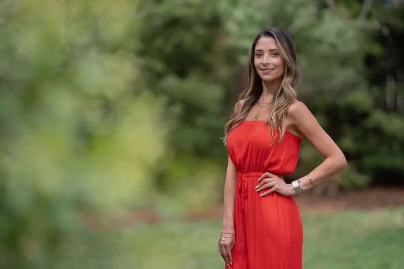Natasha poses with one hand on her hip in a red strapless dress, framed by vibrant garden landscaping.