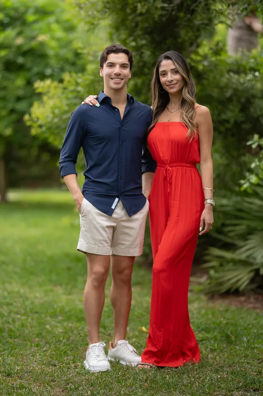 Marco and Natasha stand together on the lawn, Marco in a navy shirt and khaki shorts, Natasha in her red floor-length gown.