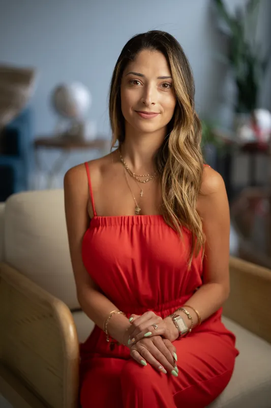 Natasha sits on a cream chair in a red strapless dress, hands folded, looking toward the camera in a modern interior space.