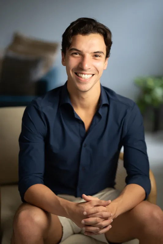 Marco grins while seated, wearing a navy button-up shirt with sleeves rolled, hands clasped in front of him indoors.