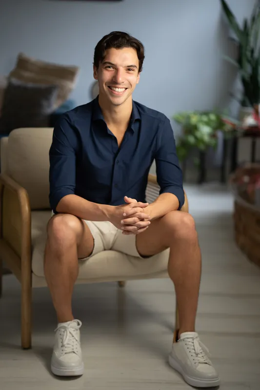 Marco sits on a cream chair in a dark blue shirt and khaki shorts, smiling at the camera with feet planted on the floor.
