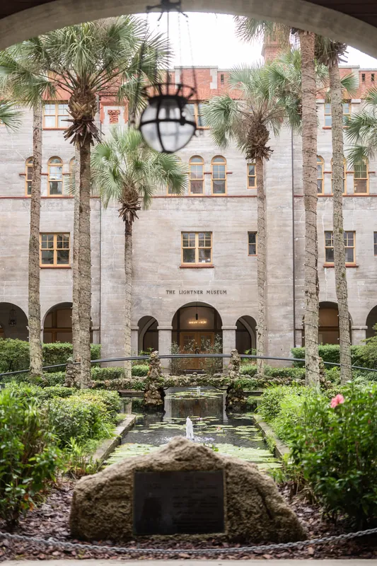 Manicured courtyard at Lightner Museum framed by a stone archway, with palm trees, fountain, and historic building facade visible beyond.