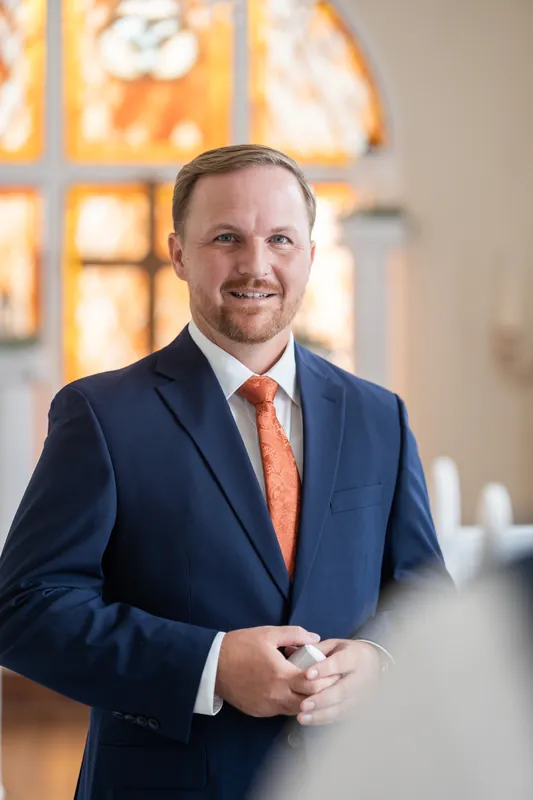 Person in navy suit and coral tie smiles at the camera inside Lightner Museum, with warm golden light from stained glass visible behind.