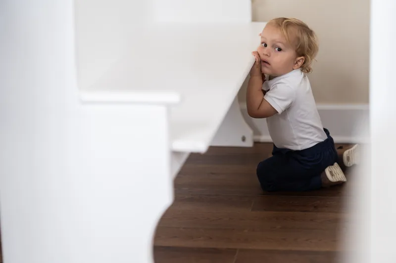 Small child in white shirt and dark pants sits alone on a wooden floor, hand to mouth, looking toward the camera.