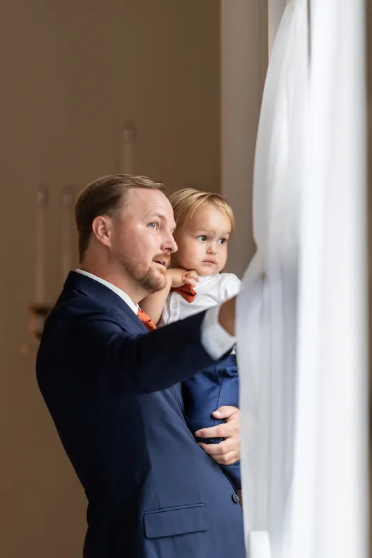 Person in dark suit holds a small child in white clothing while both look out a window at Lightner Museum.