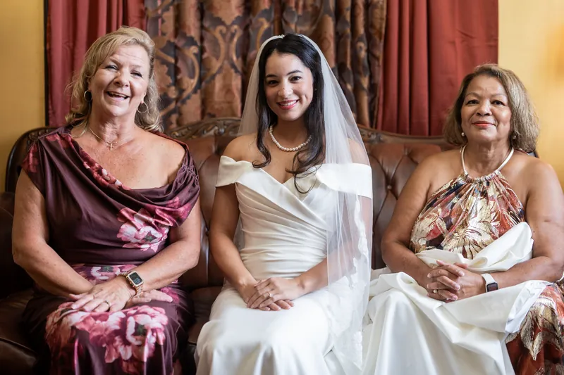 Three women smile together indoors; the center person wears a white wedding gown and veil, flanked by two guests in formal attire.