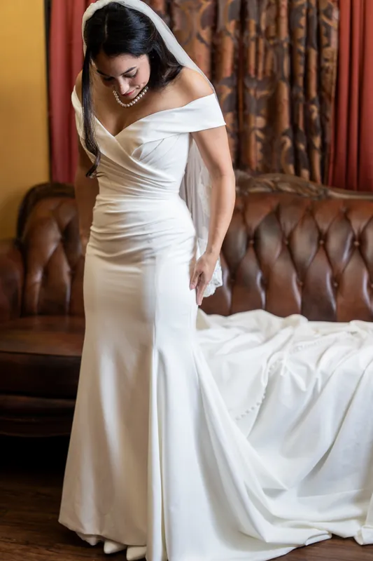 Person in a white off-shoulder gown with a veil stands beside a leather sofa at Lightner Museum, adjusting the train of their dress.