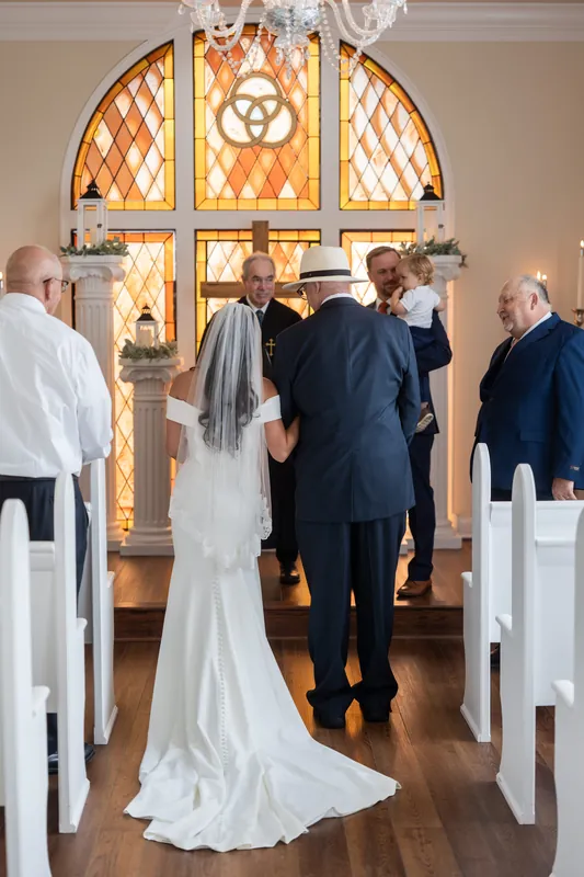 Nathan and Hillary stand at the altar facing a stained-glass window with guests seated on either side at Lightner Museum.