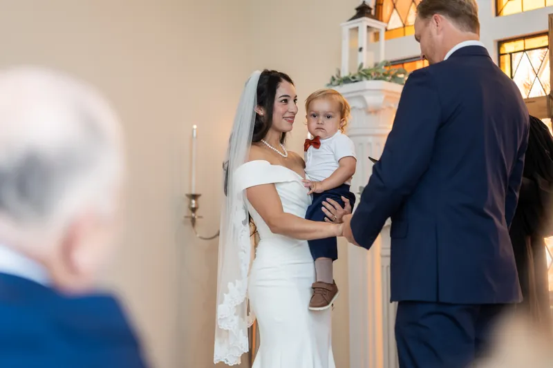 Hillary holds their young child while standing with a man in a navy suit at the altar, with ornate windows and candlelight visible behind them.