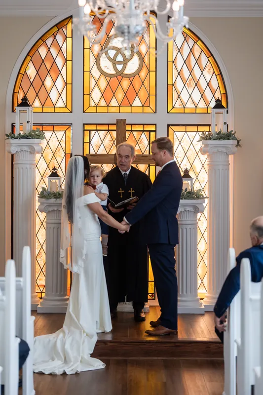 A couple exchanges vows beneath a grand arched window at Lightner Museum, with guests seated on either side in the ceremonial space.