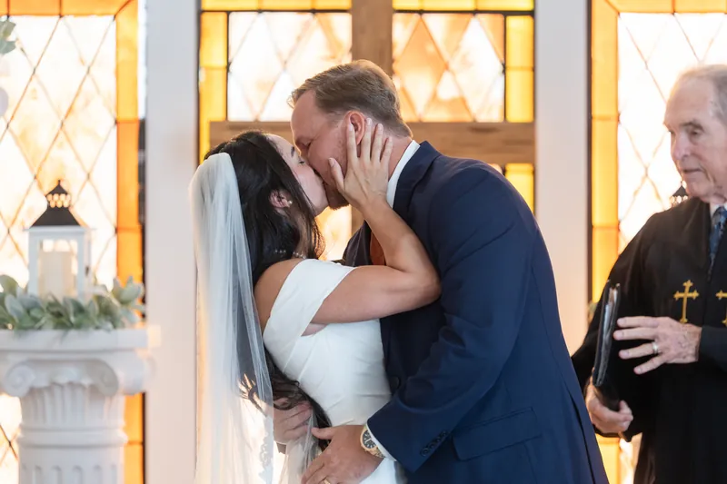 The couple shares a kiss at the altar as an officiant stands nearby, with golden light streaming through the ornate window behind them.