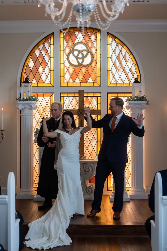 Two people in wedding attire stand before a stained-glass window at the Lightner Museum chapel during their ceremony.