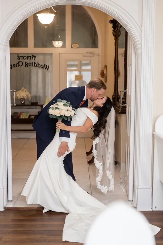 Two people in formal wear embrace in an arched doorway at Lightner Museum, one in a navy suit dipping the other in a white gown and veil.