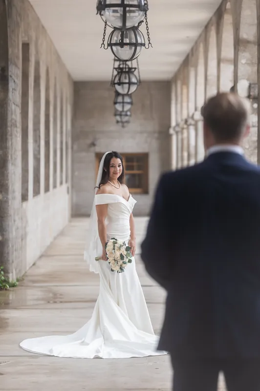 A person in an off-shoulder white gown with train stands in a colonnade hallway, facing their partner in a dark suit down the elegant corridor.