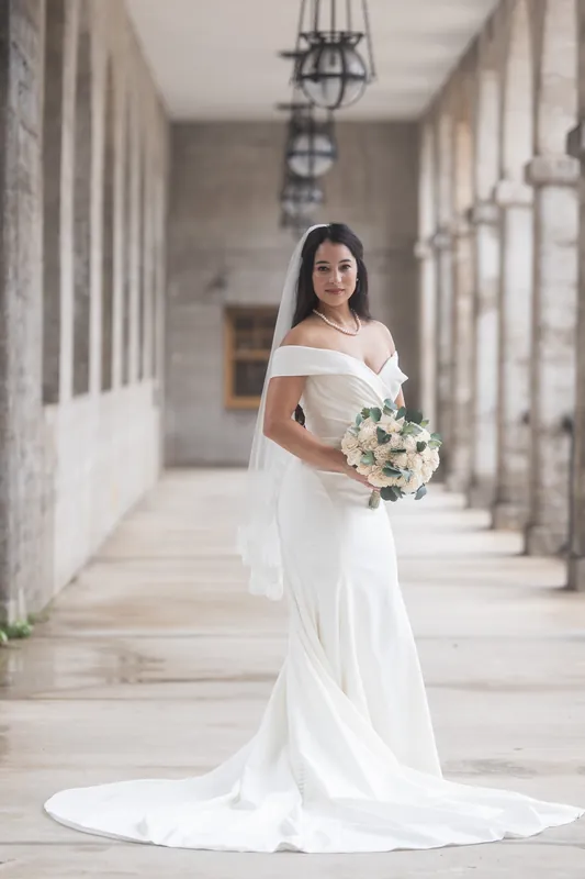 Someone in a flowing white wedding dress holds a cream and white bouquet while standing alone in Lightner Museum's arched hallway lined with columns.