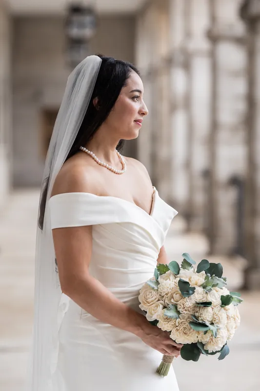 Person in white off-shoulder gown holds cream and blush bouquet with eucalyptus at Lightner Museum's colonnade.