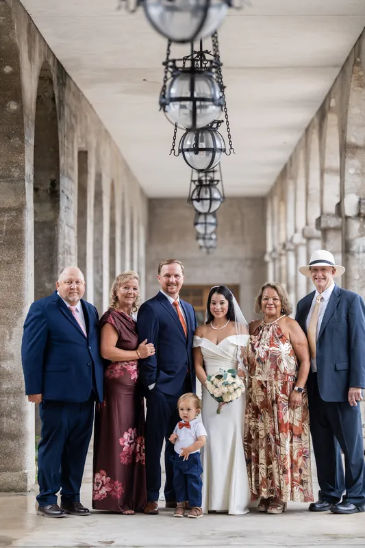 Wedding party of eight stands centered in the Lightner Museum's long colonnaded hallway beneath iron fixtures.