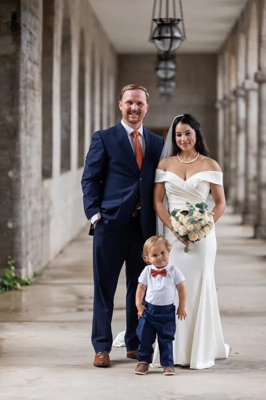 Nathan and Hillary stand with their young child in a columned corridor, Hillary in an off-shoulder gown holding a white bouquet.