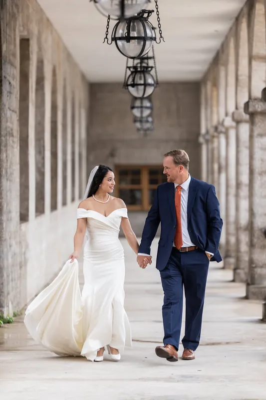 Two people in wedding attire walk hand-in-hand through a stone corridor with hanging lanterns at the Lightner Museum.