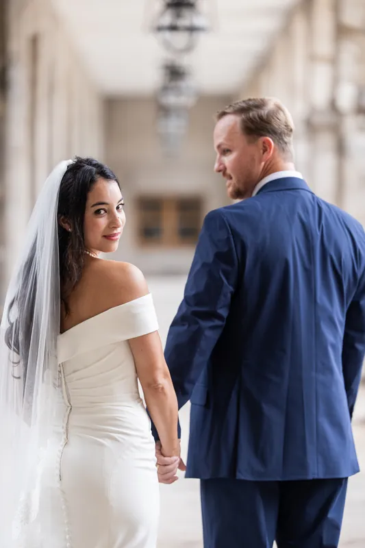 Hillary glances back over her shoulder as she walks with Nathan through an arched passageway, her veil trailing behind.