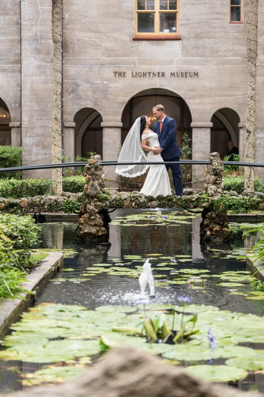 Nathan and Hillary embrace beside a lily pond in the Lightner Museum's courtyard, stone arches and manicured hedges framing them behind.