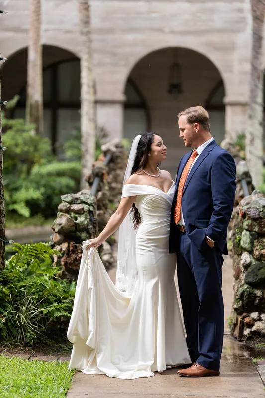 Nathan and Hillary stand close together in the Lightner Museum's garden, surrounded by lush greenery and flowering plants along a brick pathway.
