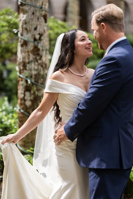 Two people in wedding attire hold hands in the Lightner Museum courtyard, surrounded by lush greenery and stone architecture.