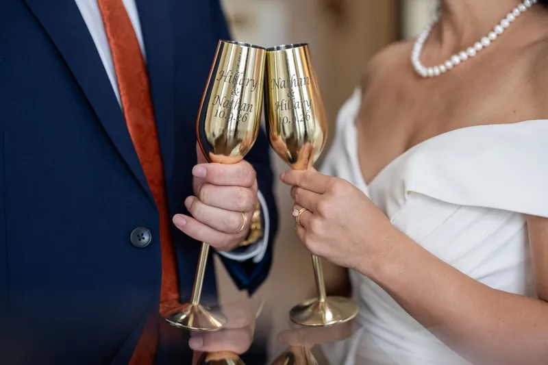 Two people in wedding attire clink champagne flutes together, their hands visible as they toast in celebration.