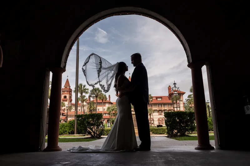 Silhouetted against the arched museum doorway, a person in a dark suit holds a veil while gazing out at the historic St. Augustine plaza beyond.