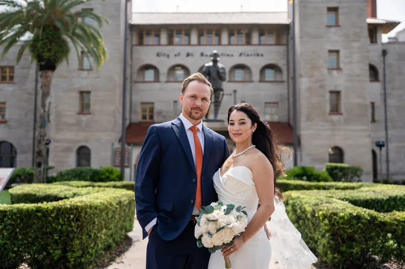 A couple stands arm-in-arm before the historic Lightner Museum facade, one holding a white bouquet in a cream gown, the other in navy.
