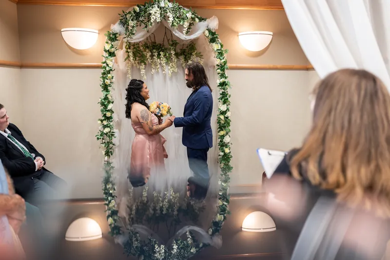 Nikki in a pink dress and Kiel in a navy suit exchange vows beneath a greenery arch during their indoor ceremony.