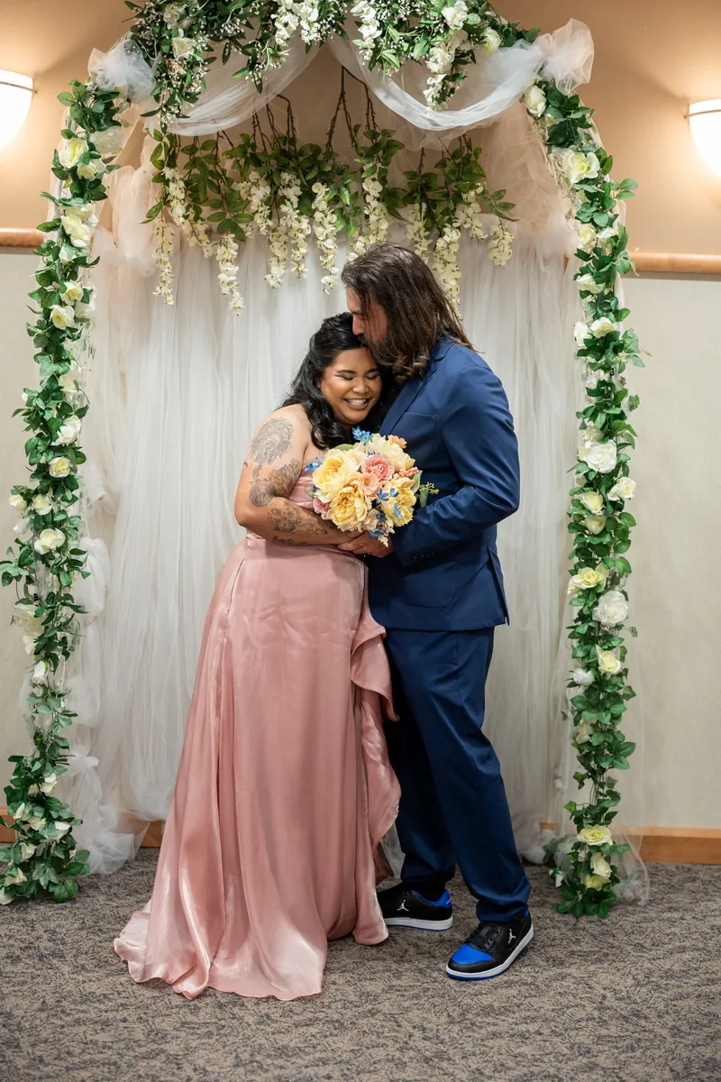 Nikki and Kiel embrace in front of the flower-framed arch, with Nikki holding her yellow and pink bouquet.