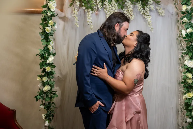 Kiel and Nikki kiss beneath the decorated arch, with Nikki's hands around his neck and shoulders.