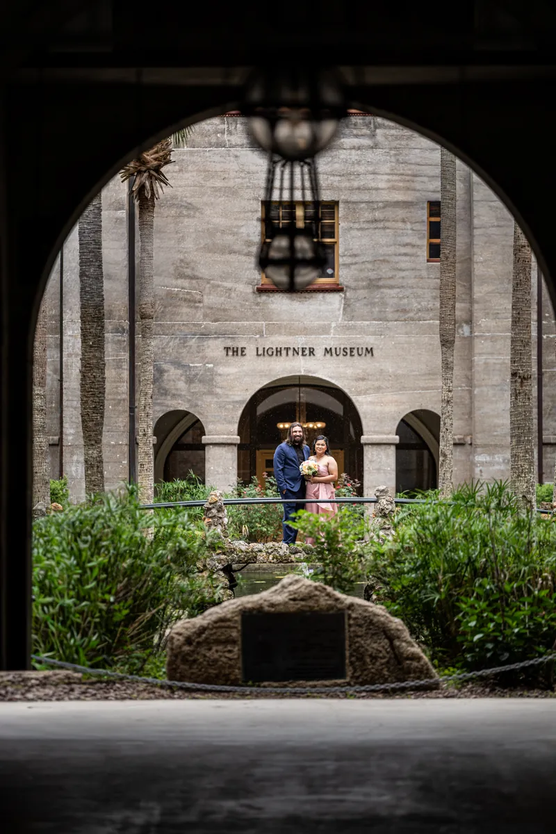 Nikki and Kiel stand together beneath an arched entrance to the Lightner Museum holding a bouquet.