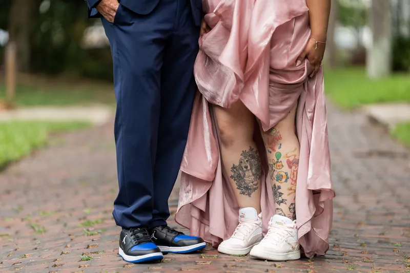 Close-up of Kiel's navy suit and blue sneakers next to Nikki's pink dress and white sneakers on a brick pathway.