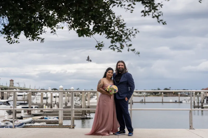 Nikki holding a bouquet and Kiel in navy stand together on a waterfront plaza overlooking boats and cityscape.