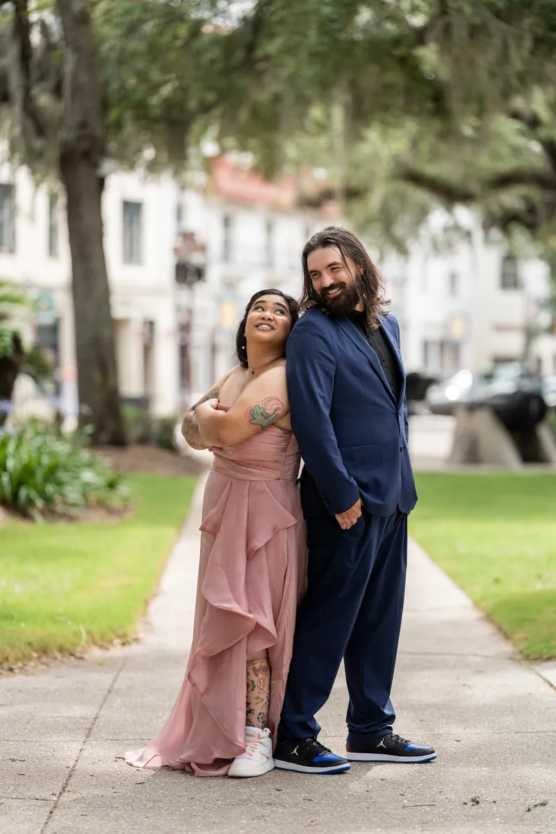 Nikki in blush and Kiel in navy pose together on a tree-lined street with historic buildings behind them.