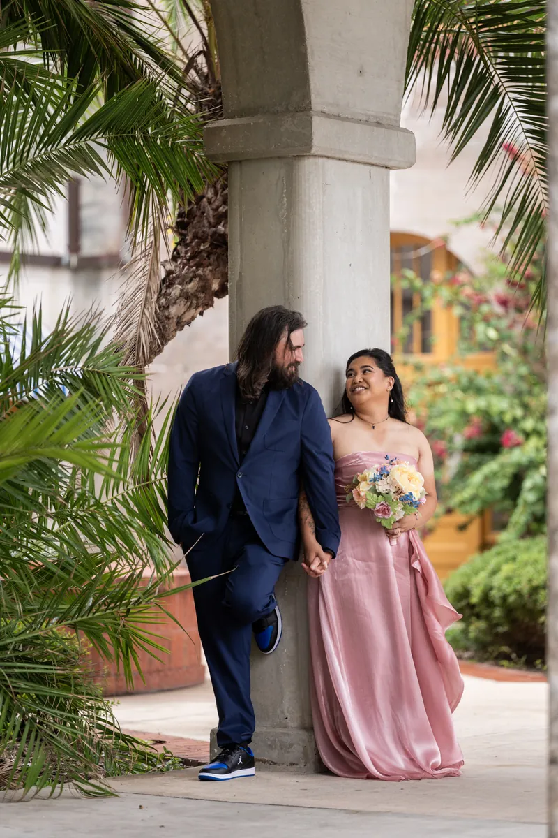 Kiel in a navy blazer and Nikki in a blush dress walk past palm tree columns on a covered loggia.