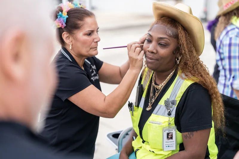 A person in a yellow safety vest and black lanyard receives face paint application from someone in dark clothing at an indoor event.