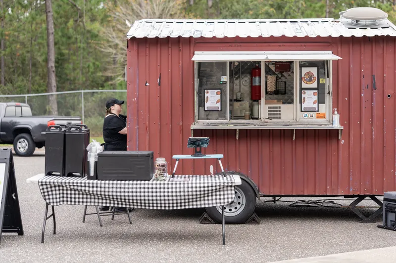 A red metal food truck with service window sits parked on pavement with checkered tablecloth and setup area nearby.